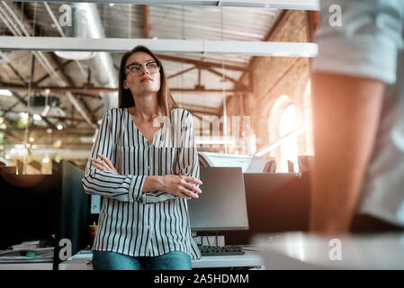 Arbeitstag. Junge Frau in Freizeitkleidung, die Arme verschränkt und Denken über etwas, während Sie die Zeit im Büro Stockfoto