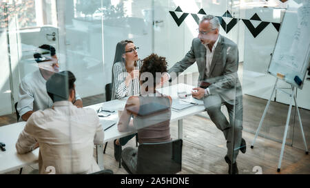 Geschäftsleute. Zuversichtlich reifer Mann in formalen Verschleiß erklärt etwas zu seinen Kollegen während der Sitzung auf dem Büro Tisch hinter der Glaswand in Stockfoto