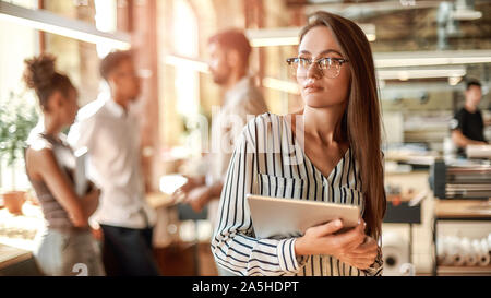 Smart und zuversichtlich. Junge Frau in Brillen Holding digital Tablet und Suchen an der Kamera, während im modernen Büro stehend Stockfoto