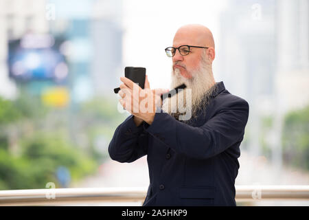 Reife bärtige kahlen Geschäftsmann bürsten Bart in der Stadt draußen Stockfoto