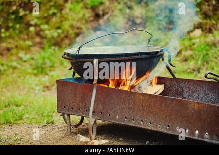 Ein metall Kessel steht auf einem Grill mit Feuer und Brennholz, in einem touristischen Parkplatz. Stockfoto