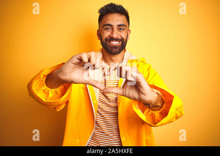 Junge indische Mann mit regenmantel stehend über isolierte gelben Hintergrund in der Liebe tut Herz Form mit Hände lächelnd. Romantische Konzept. Stockfoto