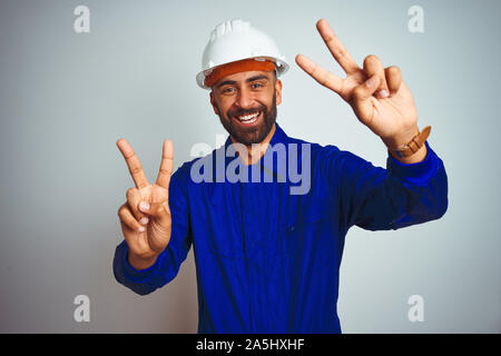 Hübscher indischer Arbeitnehmer Mann einheitliche und Helm tragen über isoliert weißer Hintergrund lächelnd in die Kamera zeigt die Finger tun Sieg unterzeichnen. Stockfoto