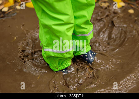 Niedrige Abschnitt eines Kindes in Grün wasserdichte Hosen und Gummistiefel in einer braunen schlammigen Pfütze stehen im Wald mit der gefallenen Blätter im Herbst an einem regnerischen Stockfoto
