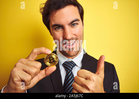 Junge gut aussehender Geschäftsmann holding Bitcoin stehend über isolierte gelber Hintergrund mit Lächeln tun ok Zeichen, Daumen hoch, mit den Fingern glücklich, excelle Stockfoto