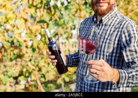 Portrait von stattlichen Winzer in seiner Hand Flasche und ein Glas Rotwein und Verkostung, Kontrolle Wein Qualität beim Stehen in der Weinberge