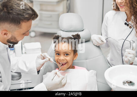 7/8-Ansicht von niedlichen afrikanischen Mädchen sitzen auf zahnmedizinischen Stuhl auf Kamera und lächelnd während Zahnärzte die Sonde und die Untersuchung der Zähne in der Klinik. Konzept der Behandlung, Gesundheit und Pflege. Stockfoto