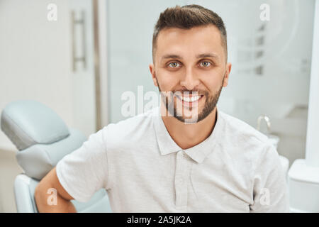 7/8-Ansicht von bärtigen fröhliche Mann Besuch Zahnarztpraxis und Härtung der Zähne. Männliche Patienten mit Kamera und lächelnd in sauberen, weißen Klinik. Konzept der Behandlung und Zahnheilkunde. Stockfoto