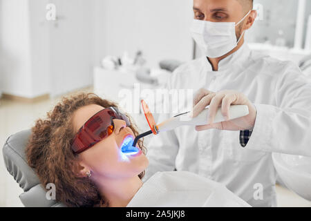 Ansicht von der Seite des männlichen Zahnarzt in weiße Uniform und Maske halten UV-und Tun in der Klinik. Junge Patientin Schutzhandschuhe liegen auf Stuhl mit geöffnetem Mund. Konzept der Pflege. Stockfoto