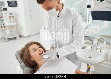 Ansicht von der Seite der hübsche junge Frau am Behandlungsstuhl und lächelnd während männliche Zahnarzt in Uniform und Maske Zähne Farbpalette. Arzt tun, die Verfahren in der Klinik. Stockfoto