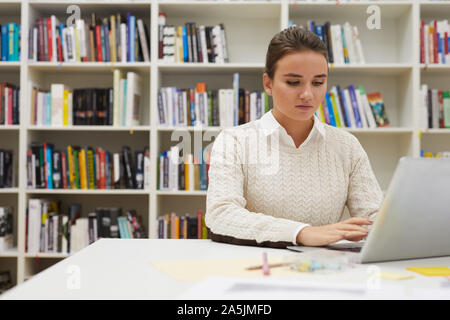 Portrait von Studentin mit Laptop während des Studiums in der Hochschule Bibliothek sitzen gegen Bücherregale, Kopie Raum Stockfoto