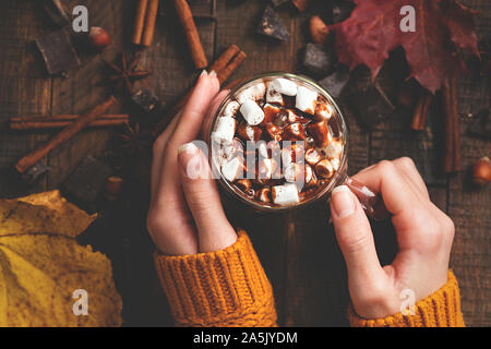 Heiße Schokolade mit Marshmallows und Schokoladensauce In weibliche Hände auf Holz- Hintergrund. Ansicht von oben. Warmen gemütlichen Getränk für Herbst oder Winter. Comfo Stockfoto