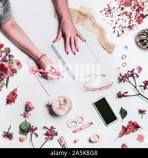 Weibliche Hände schreibt im Notebook auf weißen Desktop mit leeren Bildschirm smartphone, Tasse Kaffee, Gläser, Kosmetik, Blumen und Blätter arrangeme Stockfoto