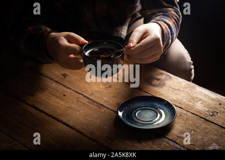 Closeup schwarzer americano Kaffee in blauer Schale in männlicher Hand, Untertasse mit Vintage Tee Löffel auf Hintergrund Holz braun vintage Dorf Tabelle. Konzept morni Stockfoto