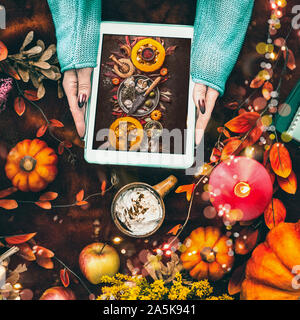 Weibliche Hände halten Tablet-PC mit Kürbisse Foto auf dunklem Hintergrund bei einer Tasse heißer Schokolade und Herbst Anordnung von Herbstlaub, brennende Kerzen Stockfoto