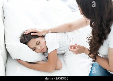 Sick cute Teen Girl im Bett zu liegen, während ihre Mutter sitzt in der Nähe und prüft die Temperatur halten Termometer in Ihrer Hand am Morgen Stockfoto