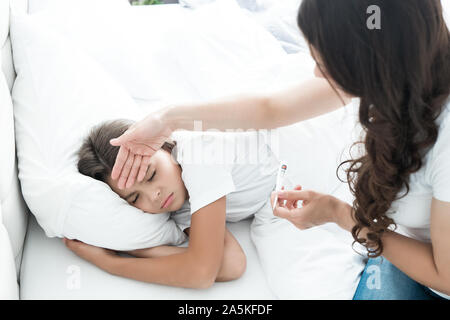Sick cute teen Mädchen liegt im Bett mit Fieber, während ihre Mutter sitzt in der Nähe und prüft die Temperatur halten Termometer in der Hand morgen Stockfoto