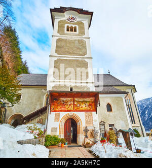 HALLSTATT, Österreich - 25. Februar 2019: Der hohe Glockenturm der Pfarrkirche mit Uhr und erhaltenen Fresken über dem Eingang, am 25. Februar in Hallstat Stockfoto