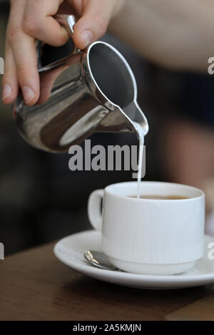 Gießen Milch/Sahne von einem silbernen Metall kann in eine frische Tasse Kaffee am Morgen. Wunderbare Art, einen Tag zu beginnen! Der Kaffee ist voller Energie! Stockfoto