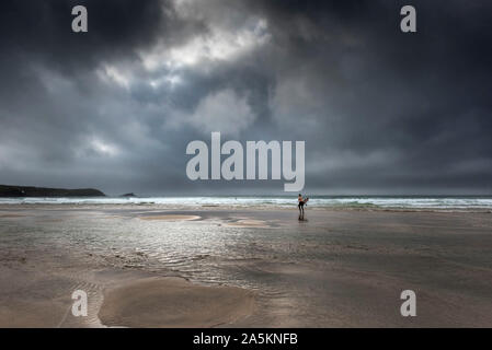 Ein Surfer, der zum Meer sein Surfbrett tragen als eine dunkle dramatische Sturm Ansätze Fistral Beach in Newquay in Cornwall. Stockfoto