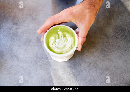 Männliche hand am tisch vegan Matcha Cappuccino mit hafermilch mit latte Kunst im Café im Freien. Stockfoto