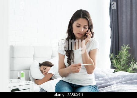 Sick cute Teen Girl schlafen im Bett, während ihre Mutter sitzt in der Nähe und ruft den Arzt holding Termometer in Ihrer Hand Stockfoto