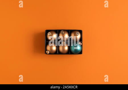 Weihnachtsbaumschmuck Zusammenhang mit gold Weihnachten Kugeln und nur eine blaue Kugel in einer schwarzen Box auf orangem Hintergrund. Oben Ansicht von Weihnachten Kugeln. Stockfoto