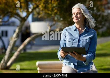 Nachdenkliche Frau mit Tablet Computer, während durch die stützmauer im Park während der sonnigen Tag stehend Stockfoto