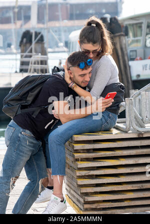 Ein junges Paar, wahrscheinlich Touristen, während der junge Mann sein Handy überprüft. In der Nähe des Canal Grande in Venedig, Italien. Stockfoto
