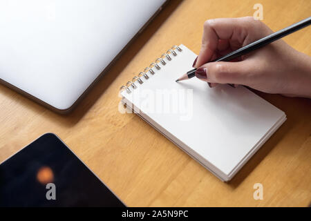 Closeup Bild der Frau hand Schreiben auf ein leeres Notebook mit Laptop, Tablet und Kaffee Tasse auf hölzernen Tisch Hintergrund Stockfoto