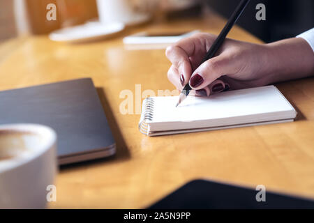 Closeup Bild der Frau hand Schreiben auf ein leeres Notebook mit Laptop, Tablet und Kaffee Tasse auf hölzernen Tisch Hintergrund Stockfoto