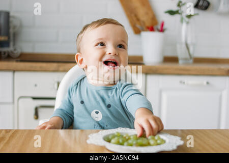 Charmantes kleines Baby boy Essen zunächst Essen Grüne Traube auf die helle Küche zu Hause Stockfoto