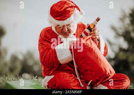 Foto der Weihnachtsmann ohne Bart, ein Bier trinken mit der roten Beutel der Geschenke in seinen Schoß. Weihnachten Stockfoto