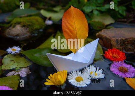 Ein Papier Boot schwimmt im dunklen Wasser, und um helle Blumen und Laub. Stockfoto