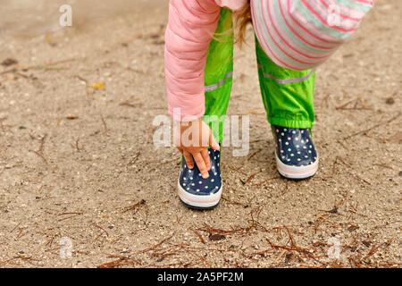 Ein Kind in Grün wasserdichte Hosen und beschmutzt, Gummistiefel, stehend auf einem nassen Feldweg in den Wald und berühren ihre Stiefel mit ihren Fingern auf ein Ra Stockfoto