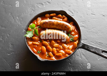 Gegrillte Würstchen mit gebackenen weißen Bohnen in Tomatensoße in der Pfanne über schwarzen Stein. Stockfoto
