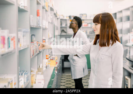 Portrait einer hübschen frau Apotheker suche Arzneimittel auf dem Regal in der Apotheke, Afrikanische männlichen Kollegen arbeiten im Hintergrund. Stockfoto