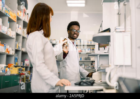 Medizin, Pharmazie, Gesundheit und Personen Konzept - glückliche afrikanischer Mann Apotheker übersicht Flasche Droge zu jungen kaukasischen Frau Apotheker an Stockfoto