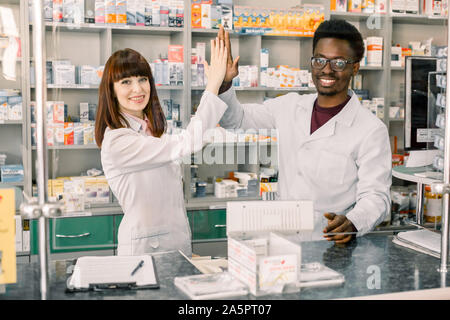 Zwei lustige gerne Kollegen Pharmazeuten, afrikanischer Mann und kaukasische Frau in der Apotheke arbeiten, lächelnd. Fünf und Spaß haben. Stockfoto