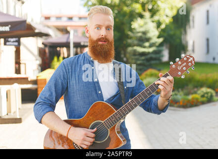 Junger bärtiger Mann trägt blaue Jeansjacke, Gitarre spielen im Freien Stockfoto