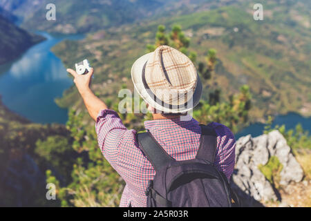 Jungen Touristen sitzen auf der Spitze des Berges, die Kamera und zeigt schöne Tal. Reisen und Natur Konzept. Stockfoto