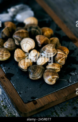 Nahaufnahme von einigen raw-Muscheln, mit crushed Eis umgeben, auf einem Holz- und schwarzem Schiefer Fach, auf einem grauen rustikalen Holztisch gelegt Stockfoto