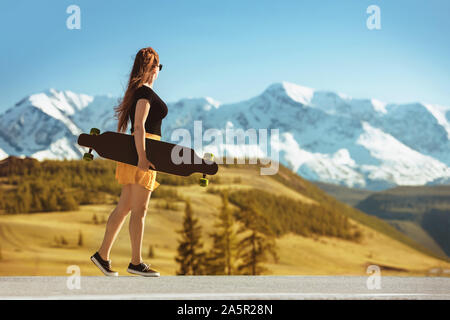 Junge Mädchen steht mit Longboard auf gerader Straße gegen Berge Stockfoto