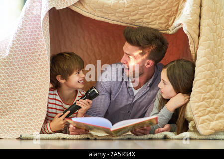 Vater und zwei Kinder lesen eine Taschenlampe mit einem Buch unter einer Decke Stockfoto