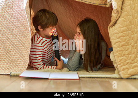 Junge ist ein Scherz mit der Taschenlampe unter eine Decke wie ein Zelt für seine Schwester Stockfoto