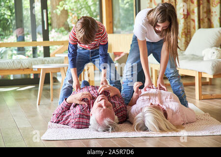 Zwei Geschwister Kinder kitzeln Oma und Opa auf dem Fußboden im Wohnzimmer Stockfoto