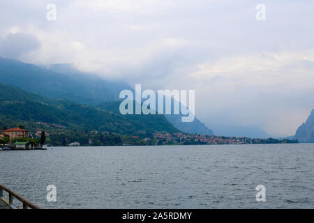 Comer See mit Gebäuden auf Küste und Alpen Berg mit Wolken im Hintergrund. Stockfoto