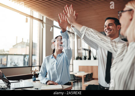 Gruppe von Lachen Mitarbeiter hohe zusammen fiving während einer Sitzung an einem Tisch in einem modernen Büro Stockfoto