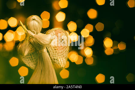 Stroh Engel betend auf einer unscharfen Weihnachten Hintergrund. Schönes Bokeh durch Weihnachtsbeleuchtung erstellt. Traditionelle handgefertigte Puppe als Weihnachtsbaum Dez Stockfoto