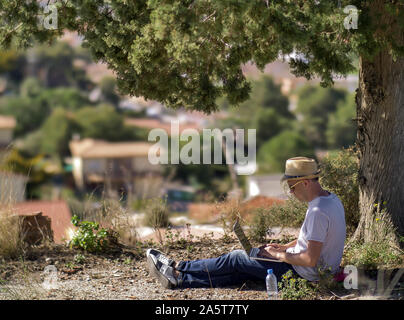 Attraktiver Kerl mit einem Laptop und in einen Hut sitzt unter einem Baum auf einem verschwommenen Hintergrund einer kleinen Stadt Stockfoto